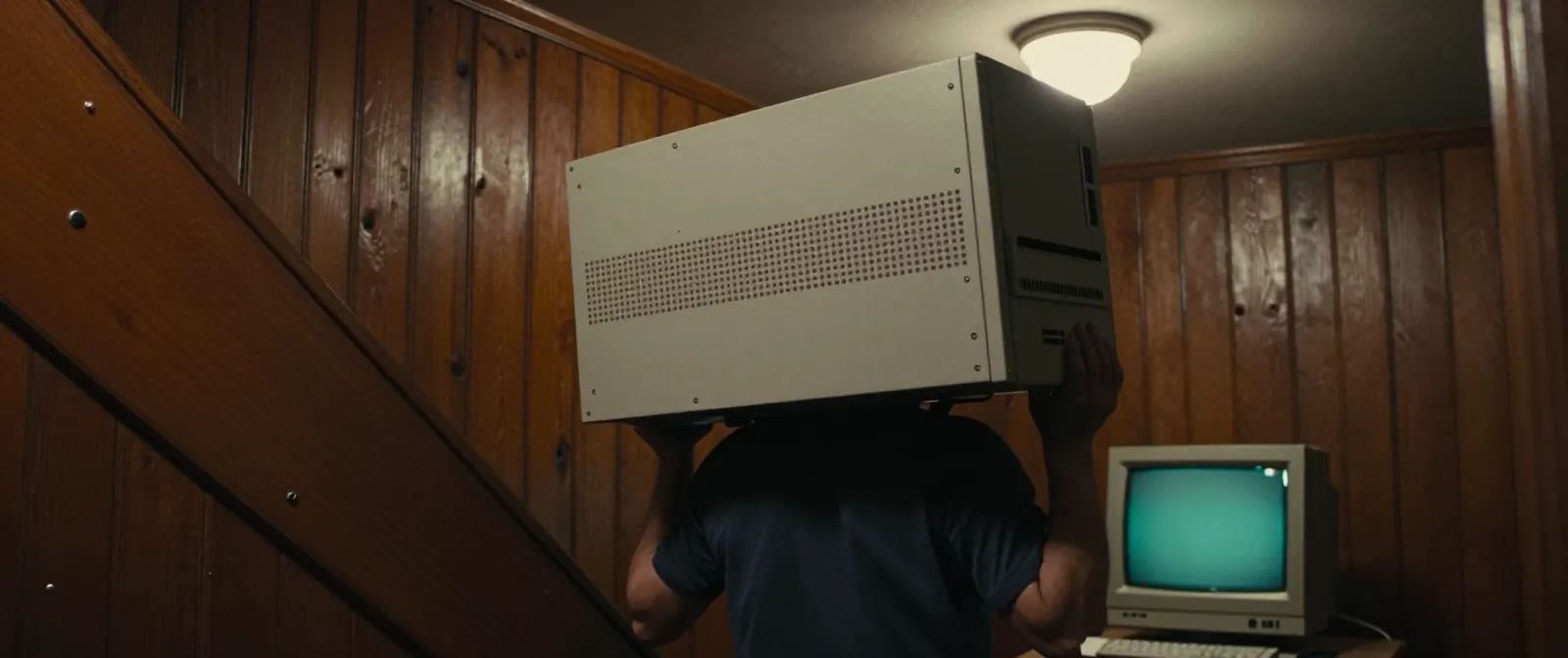 A man carries a heavy beige IBM computer case down a wood-panelled basement staircase, a monitor waiting on the desk below