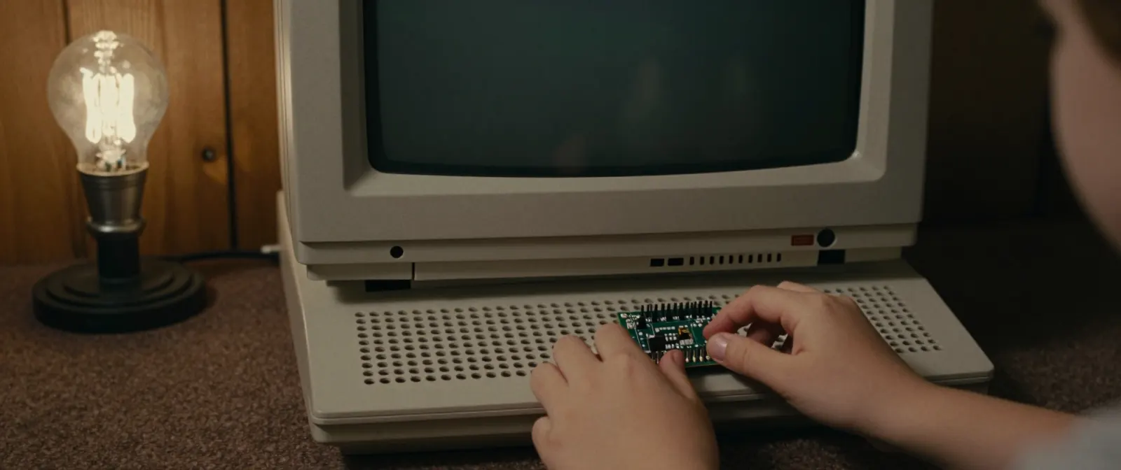 A child's hands push a small circuit board through the ventilation holes in the front grille of a beige computer